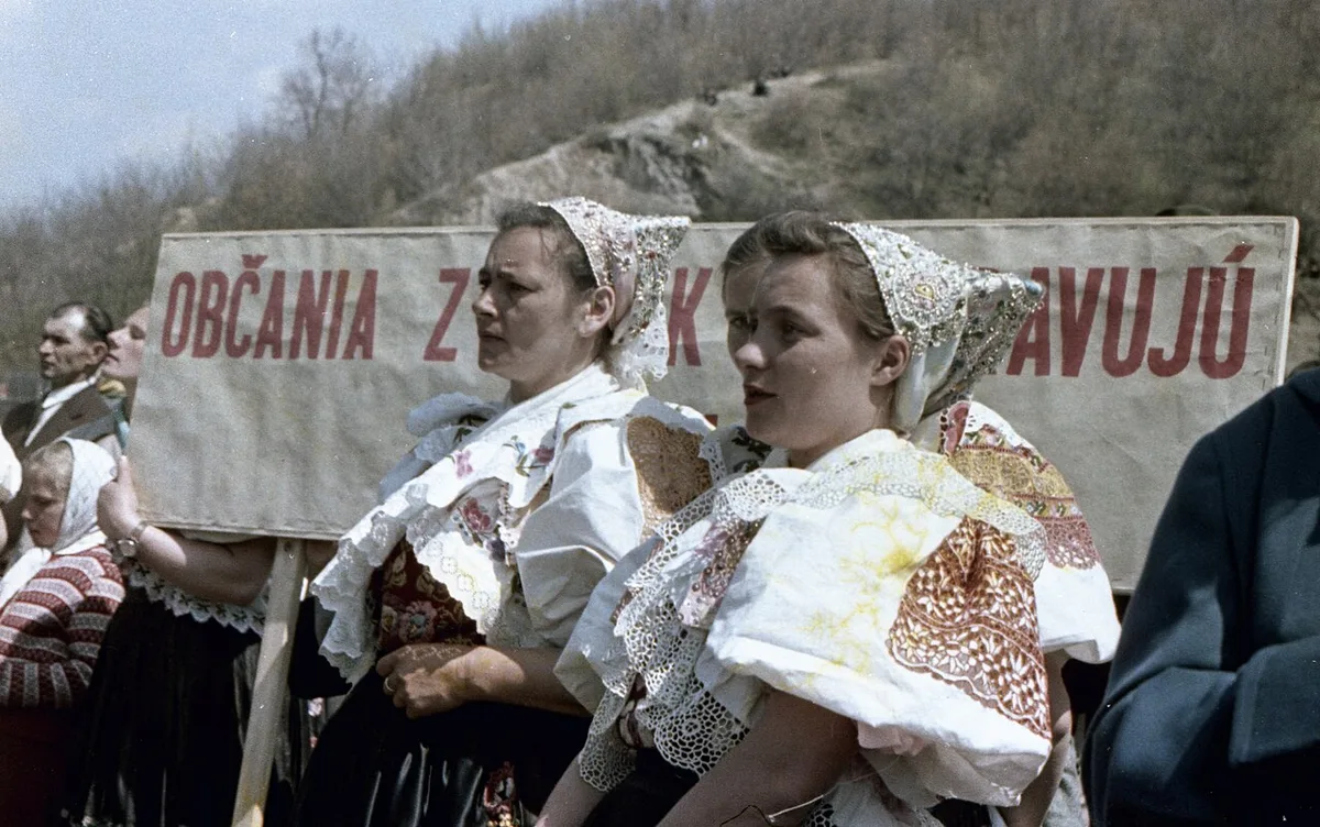 Vrouwen in geborduurde klederdracht met kanten hoofdtooi voor een spandoek met Slowaakse tekst, op een historische kleurenfoto.