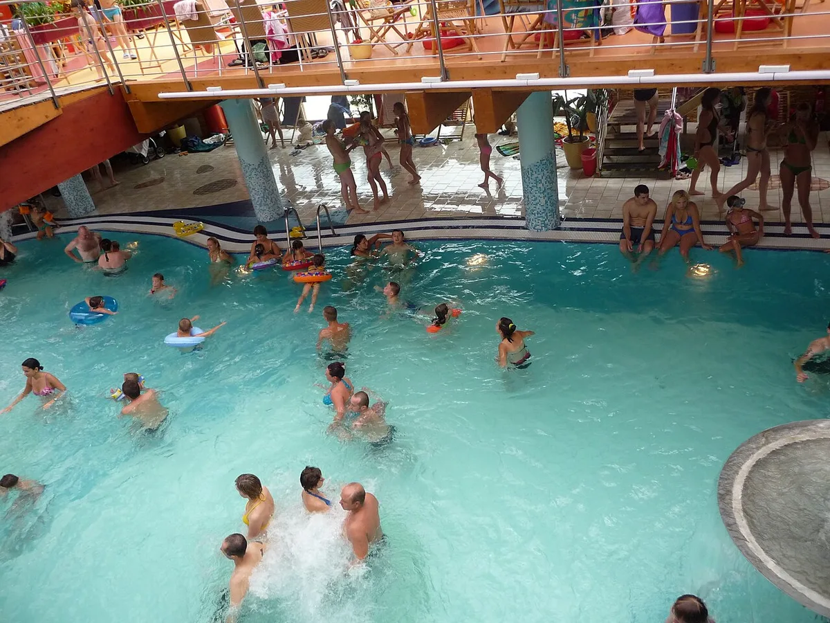 Een buitenbad met turkoois thermaal water en badgasten, met glijbanen en palmen op de achtergrond.
