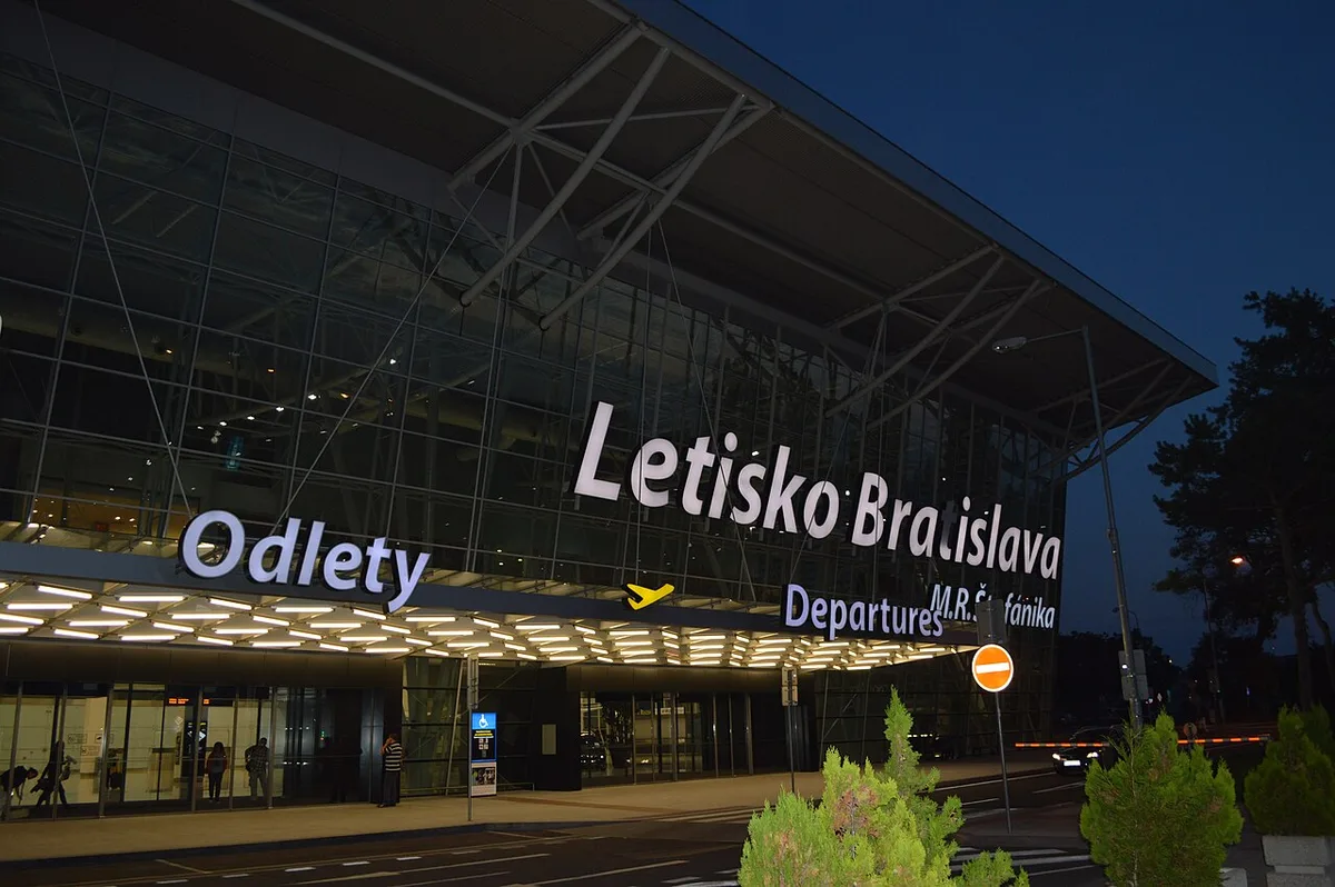 De vertrekhal van de luchthaven van Bratislava in de avondschemering, met boven de ingang de opschriften Odlety en Departures.