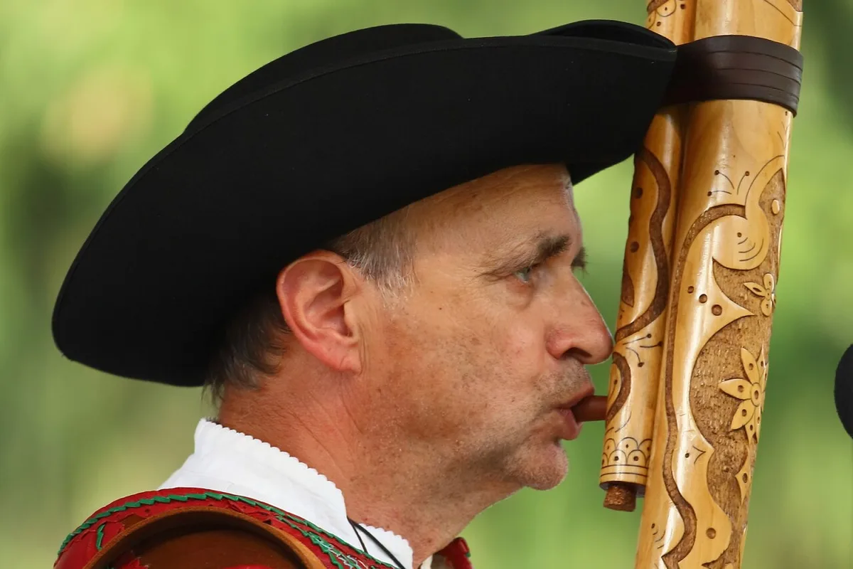 Een man met brede zwarte hoed en geborduurd vest blaast op een lange versierde fujara met een apart blaaspijpje.